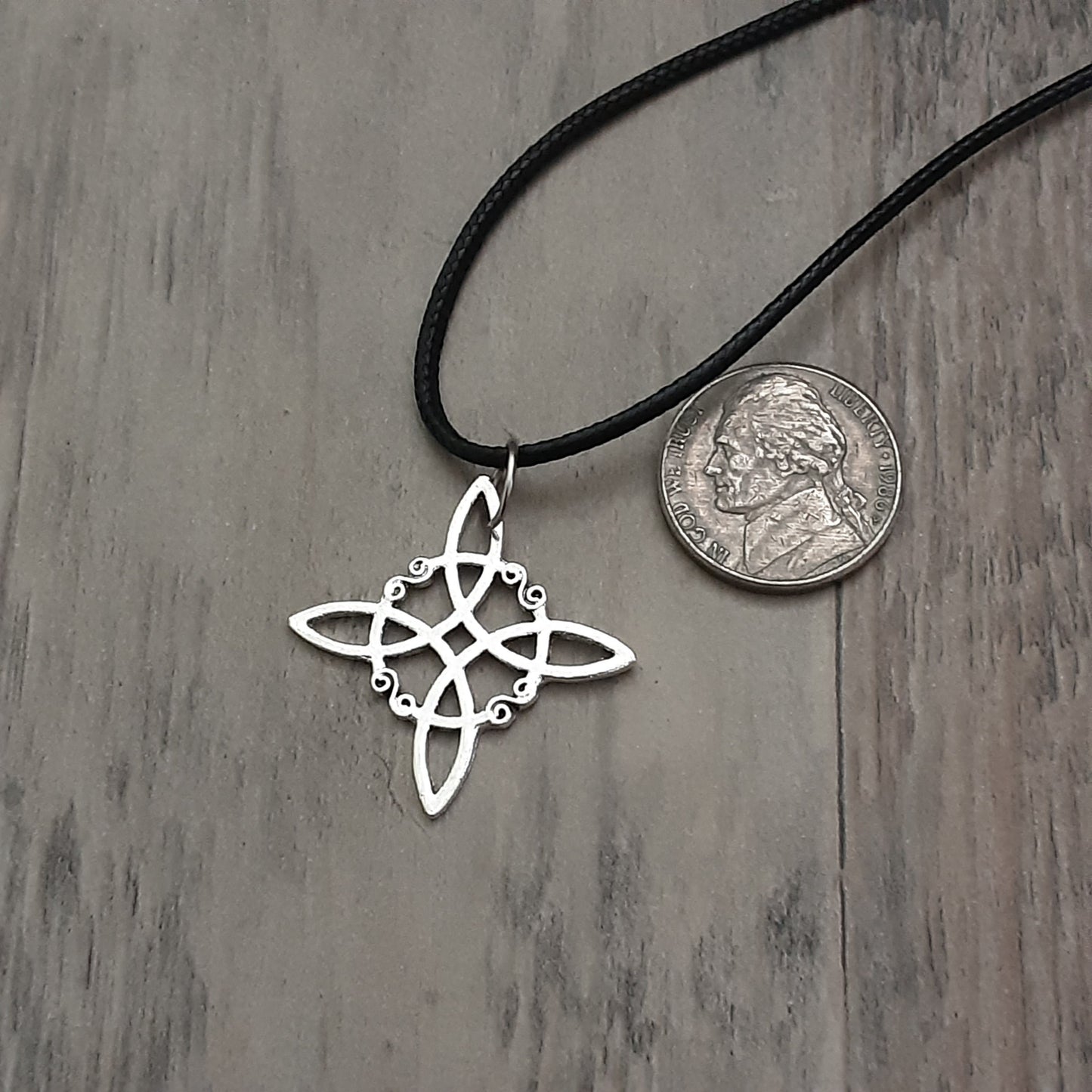 Silver witch knot symbol charm on black cord necklace next to nickel on grey wooden background